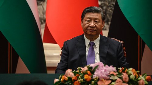 Xi Jinping sitzt vor einer Reihe von Fahnen. Vor ihm steht ein Blumenbouqet. Er schaut nachdenklich nach unten.