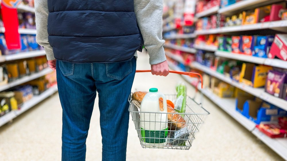 Das Bild zeigt einen Mann, der einen Einkaufskorb mit wichtigen frischen Lebensmitteln wie Brot und Milch im Supermarkt in der Hand hält.