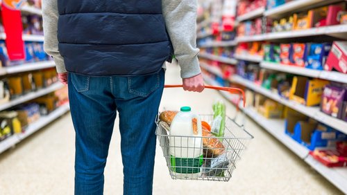 Das Bild zeigt einen Mann, der einen Einkaufskorb mit wichtigen frischen Lebensmitteln wie Brot und Milch im Supermarkt in der Hand hält.