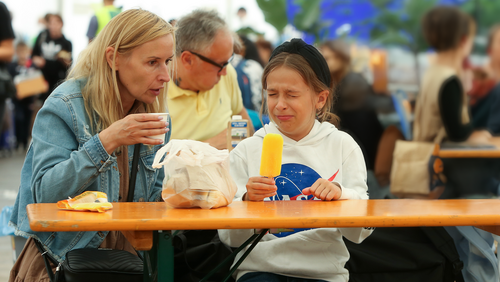 Ein Mädchen isst ein Capri-Eis und verzieht das Gesicht, ihre Mutter sitzt neben ihr